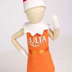 Puede incluir: Mu&ntilde;eca elfo vestida con un vestido naranja con el logo "ULTA BEAUTY". El elfo tiene un gorro rojo y blanco, guantes blancos y un cuello blanco. El fondo es blanco. La mu&ntilde;eca est&aacute; posando.