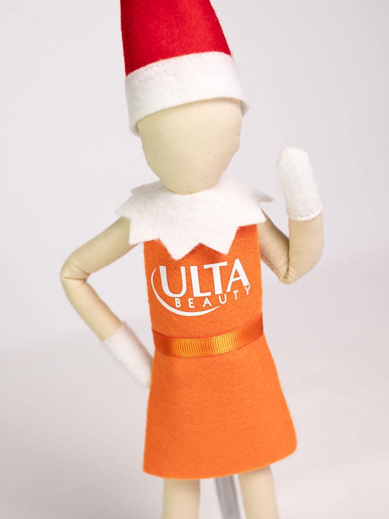 Puede incluir: Mu&ntilde;eca elfo vestida con un vestido naranja con el logo "ULTA BEAUTY". El elfo tiene un gorro rojo y blanco, guantes blancos y un cuello blanco. El fondo es blanco. La mu&ntilde;eca est&aacute; posando.