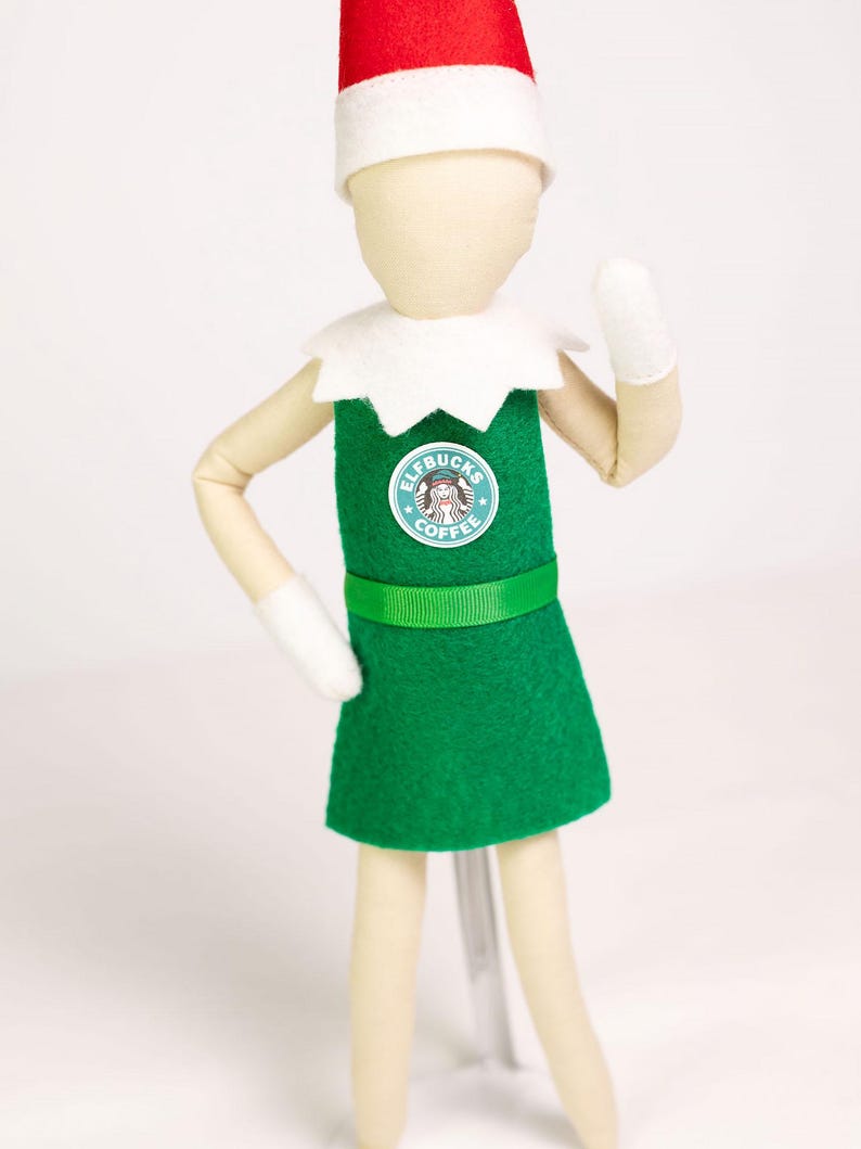May include: A fabric doll wearing a red and white pointed hat, a white collar, and a green apron featuring a circular logo that reads "ELFBUCKS COFFEE" with a central graphic. The doll has light-colored limbs, white mittens, and a plain, featureless face.
