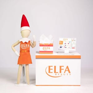 May include: An elf doll wearing an orange dress and a red hat stands next to a white counter with the text "ELFA BEAUTY". A computer monitor displays a beauty product website. A box of tissues and a keyboard are also on the counter.