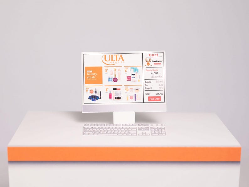 Puede incluir: Un monitor de computadora blanco que muestra un carrito de compras en l&iacute;nea de ULTA Beauty. La pantalla muestra varios productos de belleza y un resumen del carrito. Un teclado blanco est&aacute; debajo del monitor, y el escritorio tiene un acento naranja.
