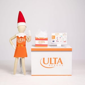 May include: An orange and white display featuring the ULTA Beauty logo, a computer screen, and a tissue box. A wooden doll wearing an orange dress and a red elf hat stands next to the display. The computer screen displays a website.