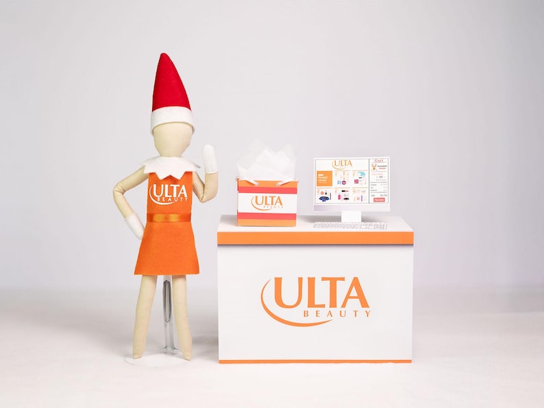 Puede incluir: Una exhibici&oacute;n naranja y blanca con el logotipo de ULTA Beauty, una pantalla de computadora y una caja de pa&ntilde;uelos. Una mu&ntilde;eca de madera con un vestido naranja y un gorro de elfo rojo est&aacute; junto a la exhibici&oacute;n. La pantalla muestra un sitio web.