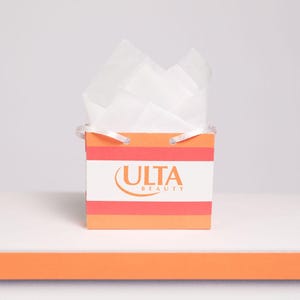 Puede incluir: Una peque&ntilde;a bolsa de regalo con el logotipo de Ulta Beauty, con rayas naranjas y blancas. La bolsa est&aacute; llena de papel de seda blanco y tiene asas blancas peque&ntilde;as. La bolsa est&aacute; sobre una superficie blanca con una franja naranja.