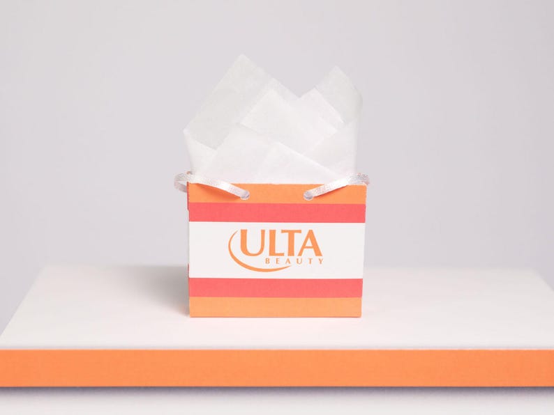 Puede incluir: Una peque&ntilde;a bolsa de regalo con el logotipo de Ulta Beauty, con rayas naranjas y blancas. La bolsa est&aacute; llena de papel de seda blanco y tiene asas blancas peque&ntilde;as. La bolsa est&aacute; sobre una superficie blanca con una franja naranja.