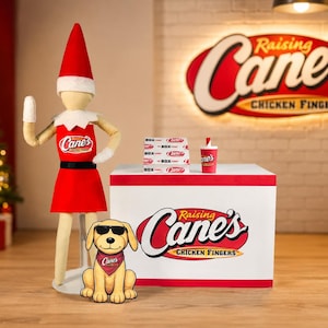 May include: A doll wearing a red and white elf hat and a Raising Cane's Chicken Fingers uniform stands next to a white counter. A plush dog with sunglasses and a red bandana sits in front of a stack of food boxes and a drink cup with the logo.