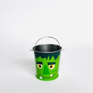 May include: A green metal bucket with a silver handle, featuring a cartoon Frankenstein face. The bucket has yellow eyes, a stitched mouth, and dark green hair. Perfect for Halloween treats or decorations.