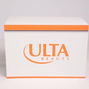 Puede incluir: Una caja blanca con ribetes naranjas y las palabras "ULTA BEAUTY" en naranja. La caja es cuadrada y parece estar hecha de cart&oacute;n o un material similar. El logotipo est&aacute; centrado en la parte frontal de la caja.