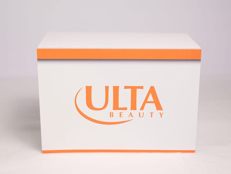 Puede incluir: Una caja blanca con ribetes naranjas y las palabras "ULTA BEAUTY" en naranja. La caja es cuadrada y parece estar hecha de cart&oacute;n o un material similar. El logotipo est&aacute; centrado en la parte frontal de la caja.