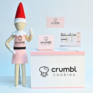 May include: An elf figurine wearing a pink dress and a red hat stands next to a white counter with the "crumbl cookies" logo. A computer screen displays a cart page. Additional "crumbl cookies" branding is visible.