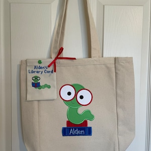 May include: A beige canvas tote bag with red handles. The bag has a green worm with red glasses and a blue book that says "Alden" on it. A smaller beige pouch with a green background says "Alden's Library Card" with a cartoon worm reading a book.