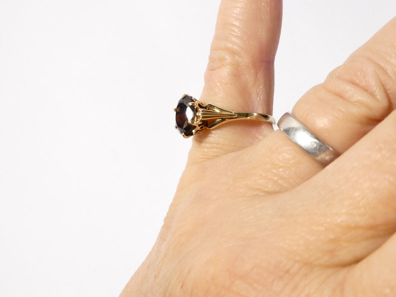 Op de afbeelding: Een gouden ring met een grote, donkerbruine edelsteen in een kraalbezetting. De ring wordt aan een vinger gedragen.
