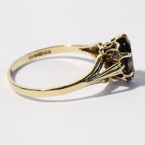 Op de afbeelding: Een gouden ring met een grote, ovale, donkerbruine edelsteen gezet in een delicate, sierlijke setting.