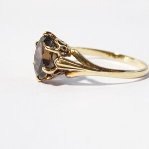 Op de afbeelding: Een gouden ring met een grote, ovale, bruine edelsteen gezet in een decoratieve, sierlijke zetting.
