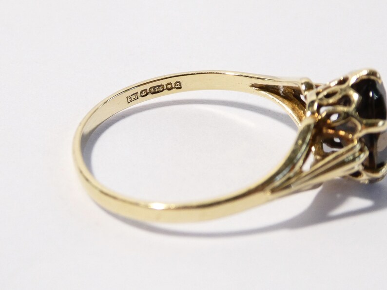 Op de afbeelding: Een gouden ring met een grote, donkerbruine edelsteen gezet in een delicaat, sierlijk ontwerp. De ring is gemarkeerd met "375" aan de binnenkant van de band.