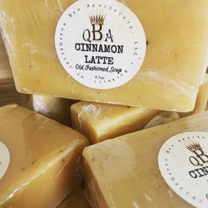 May include: A stack of four bars of handmade soap wrapped in plastic. The top bar has a label that reads "QBA Cinnamon Latte Old Fashioned Soap 4.7 oz".