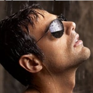 May include: A man wearing sunglasses with raindrops on the lenses, looking up at the sky.
