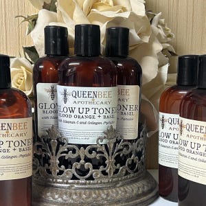 May include: Several brown glass bottles with black caps are displayed in a decorative metal holder. The bottles have white labels with the text "QUEENBEE APOTHECARY GLOW UP TONER" and ingredient information. The background features white flowers.