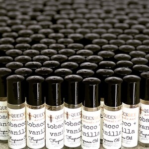 Może przedstawiać: Widok z bliska na wiele małych szklanych butelek z czarnymi nakrętkami typu roll-on. Butelki są oznaczone tekstem "Queen Bee Apothecary" i "Tobacco + Vanilla" białym fontem. Butelki są ułożone w rzędach na powierzchni.