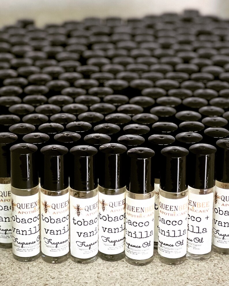 Może przedstawiać: Widok z bliska na wiele małych szklanych butelek z czarnymi nakrętkami typu roll-on. Butelki są oznaczone tekstem "Queen Bee Apothecary" i "Tobacco + Vanilla" białym fontem. Butelki są ułożone w rzędach na powierzchni.