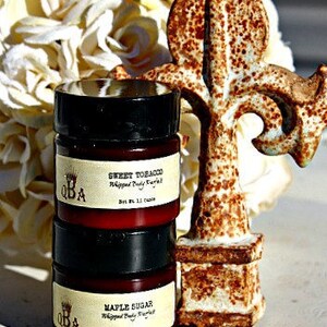 May include: Two small jars of whipped body butter, one labeled "Sweet Tobacco" and the other "Maple Sugar". The jars are sitting on a white surface next to a rusty metal decorative piece.