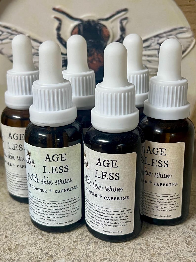 May include: A collection of six brown glass bottles with white droppers. Each bottle has a label that reads "AGE LESS peptide skin serum with copper + caffeine". The bottles are arranged in a group.