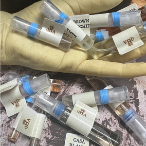 May include: A collection of small glass perfume sample bottles with blue and white labels. The labels have the text "QBA" and "Casa Blanca" with a crown logo. The bottles are arranged on a brown surface.