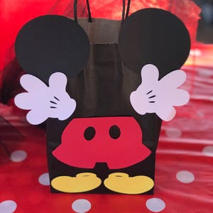 Może przedstawiać: Czarna papierowa torba prezentowa ozdobiona uszami Myszki Miki, rękami, spodenkami i butami. Torba ma czerwono-białe kropki w tle.