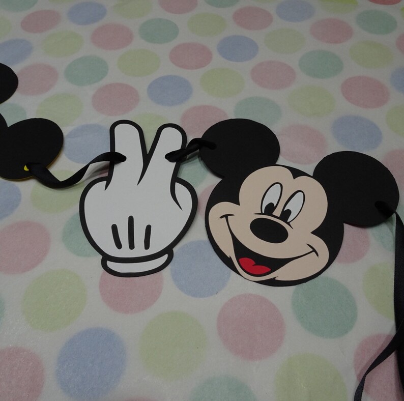 May include: A Mickey Mouse-themed banner featuring a smiling Mickey Mouse face, a white gloved hand making a peace sign, and black Mickey Mouse ears. The banner is connected by black ribbons and is set against a polka dot background.