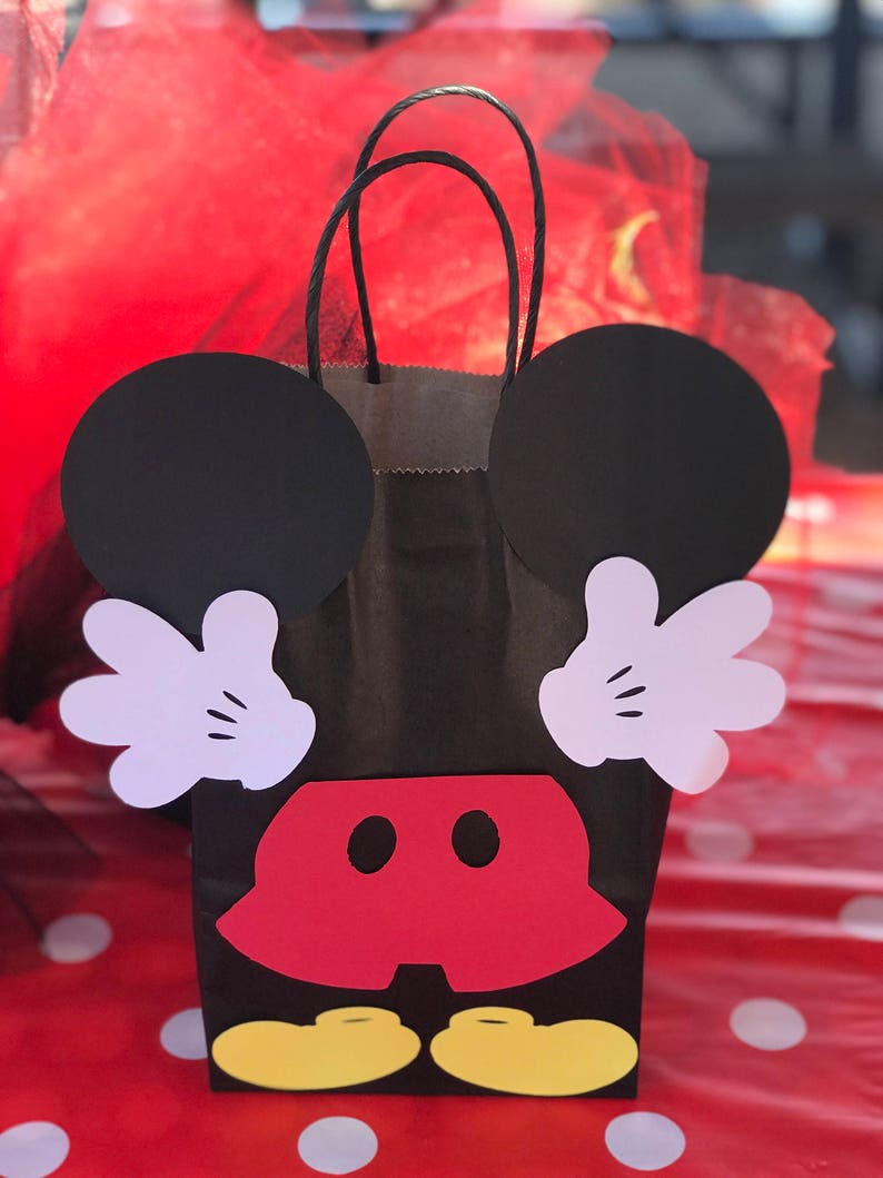 Może przedstawiać: Czarna papierowa torba prezentowa ozdobiona uszami Myszki Miki, rękami, spodenkami i butami. Torba ma czerwono-białe kropki w tle.