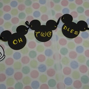 May include: A black Mickey Mouse-themed banner with the words "OH TWO DLES" in yellow. The banner includes Mickey Mouse heads and white gloved hands. The background is a polka dot pattern.