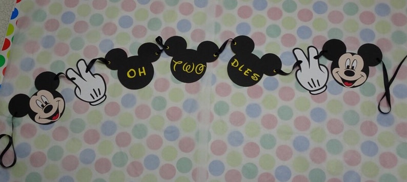 May include: A black Mickey Mouse-themed banner with the words "OH TWO DLES" in gold lettering. The banner includes Mickey Mouse heads and white gloved hands. The background is a polka dot pattern in various pastel colors.