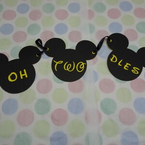 May include: A black banner with three Mickey Mouse head cutouts. The words "OH TWO DLES" are written in yellow script on the cutouts. The banner is strung with black ribbon and is set against a polka dot background.