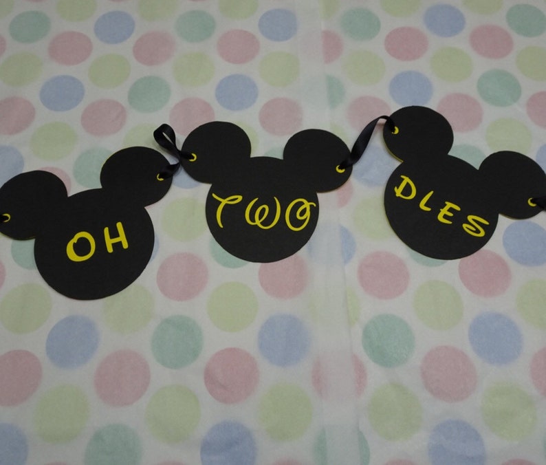 May include: A black banner with three Mickey Mouse head cutouts. The words "OH TWO DLES" are written in yellow script on the cutouts. The banner is strung with black ribbon and is set against a polka dot background.