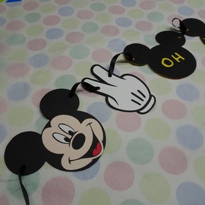 May include: A garland featuring Mickey Mouse's face, a white gloved hand, and Mickey Mouse ears with the letters "OH" in yellow. The garland is made of black and white cardstock with black ribbon for hanging. The background is a polka dot pattern.