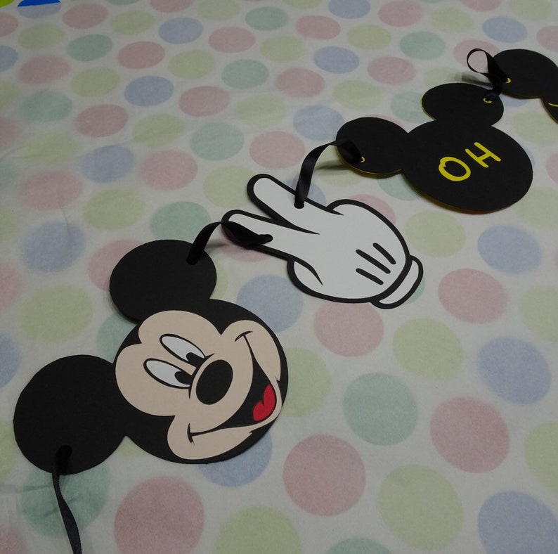 May include: A garland featuring Mickey Mouse's face, a white gloved hand, and Mickey Mouse ears with the letters "OH" in yellow. The garland is made of black and white cardstock with black ribbon for hanging. The background is a polka dot pattern.