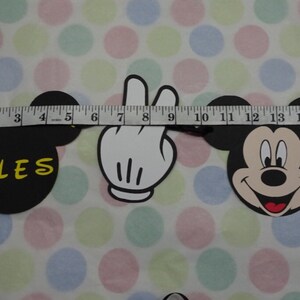 May include: A black Mickey Mouse-themed banner with a white hand and the word "DLES" in yellow. The banner is measured with a tape measure. The background is a white fabric with colorful polka dots.