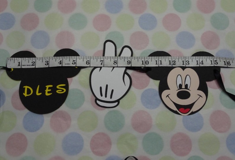 May include: A black Mickey Mouse-themed banner with a white hand and the word "DLES" in yellow. The banner is measured with a tape measure. The background is a white fabric with colorful polka dots.