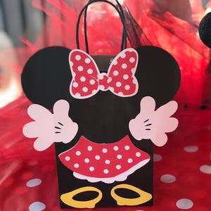 Może przedstawiać: Czarna i czerwona torba na prezenty na imprezę w stylu Minnie Mouse z białymi kropkami. Torba ma uchwyt i ozdobiona jest kokardką, sp&oacute;dnicą i butami.