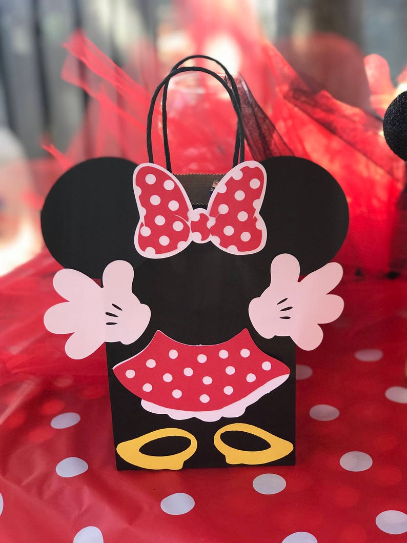 Może przedstawiać: Czarna i czerwona torba na prezenty na imprezę w stylu Minnie Mouse z białymi kropkami. Torba ma uchwyt i ozdobiona jest kokardką, sp&oacute;dnicą i butami.