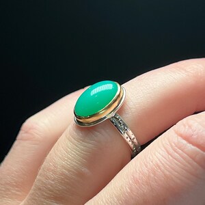 May include: A silver ring with a gold bezel holding a large, oval, bright green gemstone. The ring band has a textured design. The ring is shown on a person's finger against a dark background.