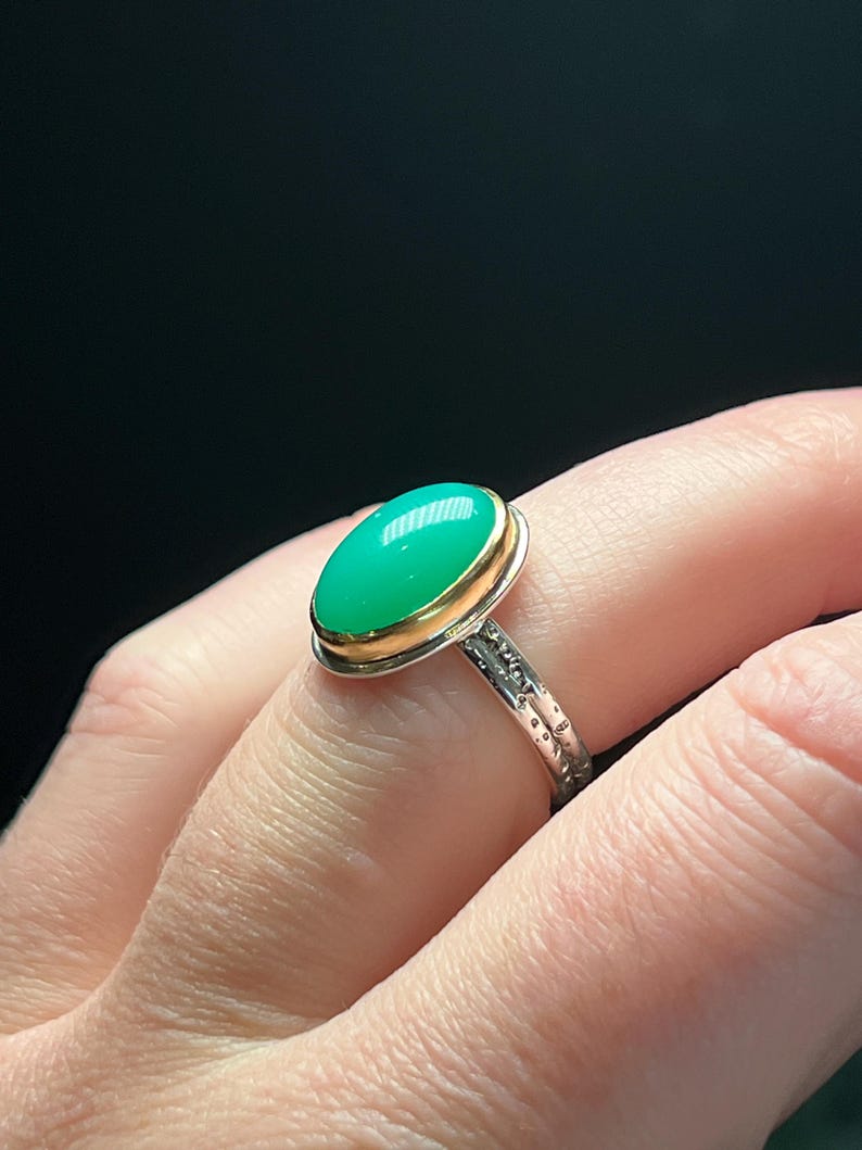 May include: A silver ring with a gold bezel holding a large, oval, bright green gemstone. The ring band has a textured design. The ring is shown on a person's finger against a dark background.