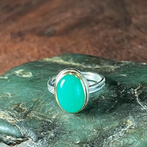 May include: A silver ring with a gold band and a large, oval, bright green gemstone. The ring is set on a textured, dark green surface, with a blurred brown background.