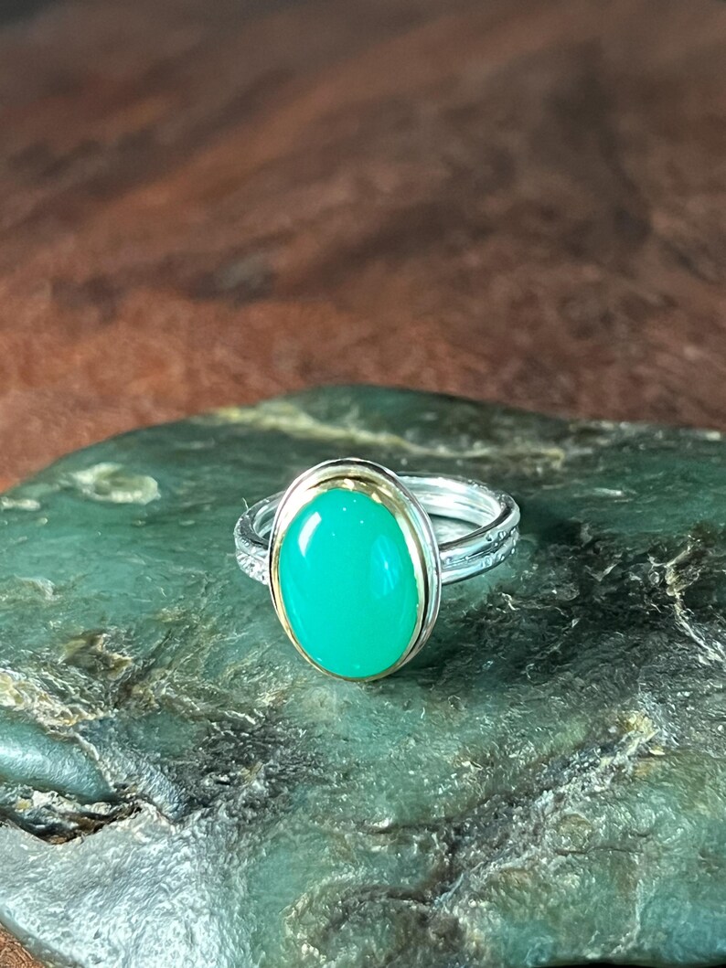 May include: A silver ring with a gold band and a large, oval, bright green gemstone. The ring is set on a textured, dark green surface, with a blurred brown background.