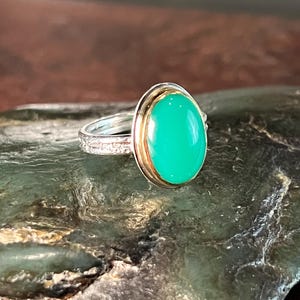 May include: A silver ring with a gold bezel setting a large, oval, bright green gemstone. The ring band has a detailed, textured design. The ring is displayed on a dark, textured surface.