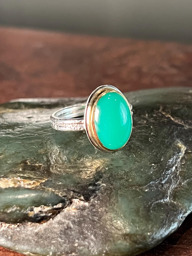 May include: A silver ring with a gold bezel setting a large, oval, bright green gemstone. The ring band has a detailed, textured design. The ring is displayed on a dark, textured surface.