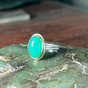 May include: A silver ring with a bright green oval gemstone set in a gold bezel. The ring is displayed on a green stone surface, showcasing the contrast between the metal and the gemstone.