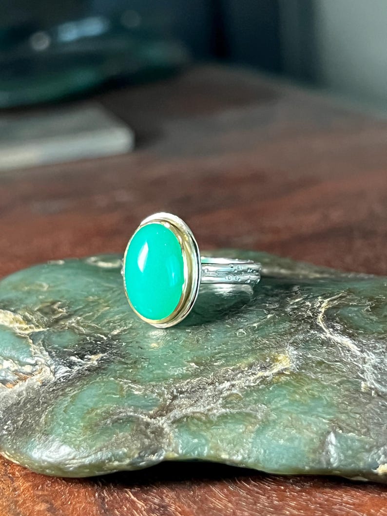 May include: A silver ring with a bright green oval gemstone set in a gold bezel. The ring is displayed on a green stone surface, showcasing the contrast between the metal and the gemstone.