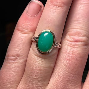 May include: A silver and gold ring featuring a large, oval-shaped, bright green gemstone. The gemstone is set in a gold bezel, and the ring band is silver with delicate detailing. The ring is shown on a finger.