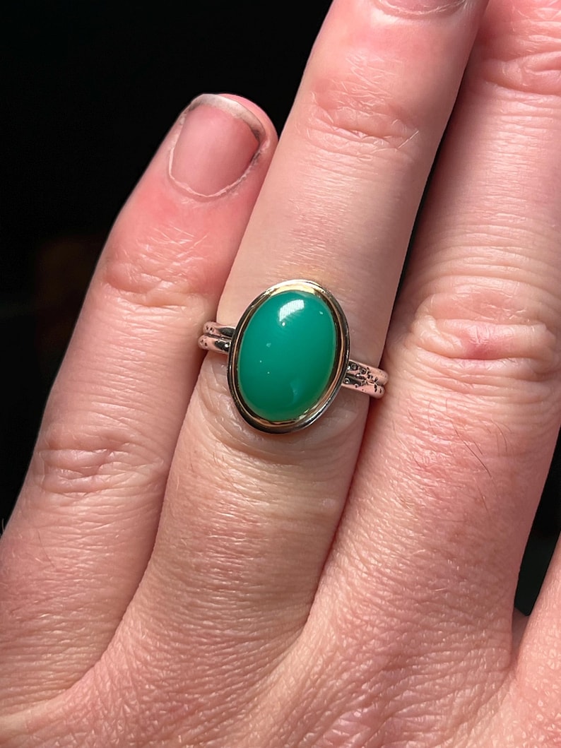 May include: A silver and gold ring featuring a large, oval-shaped, bright green gemstone. The gemstone is set in a gold bezel, and the ring band is silver with delicate detailing. The ring is shown on a finger.